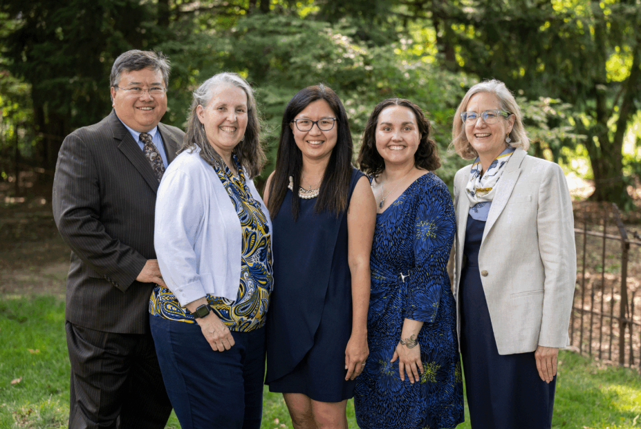 Five people stand in a row, smiling at the camera, standing in front of a wooded area outdoors.