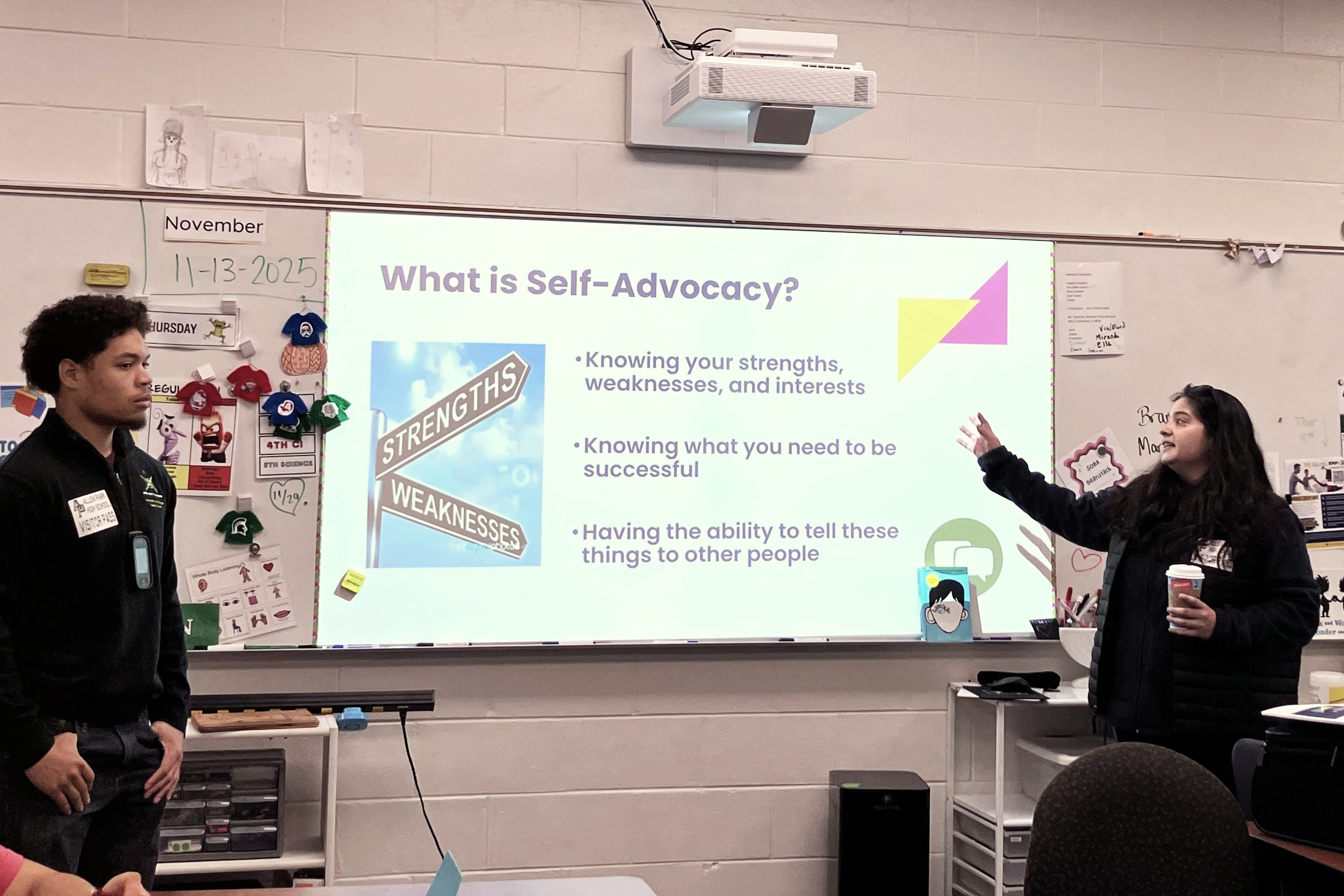 Two teachers stand in front of a classroom, referring to text projected onto the whiteboard.